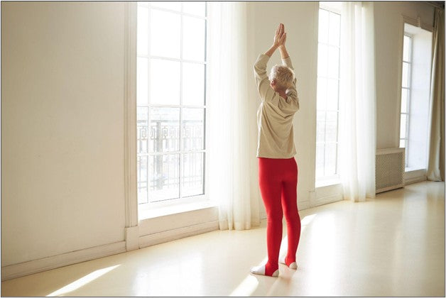 An older woman stretching with her hands overhead in a sunlit room