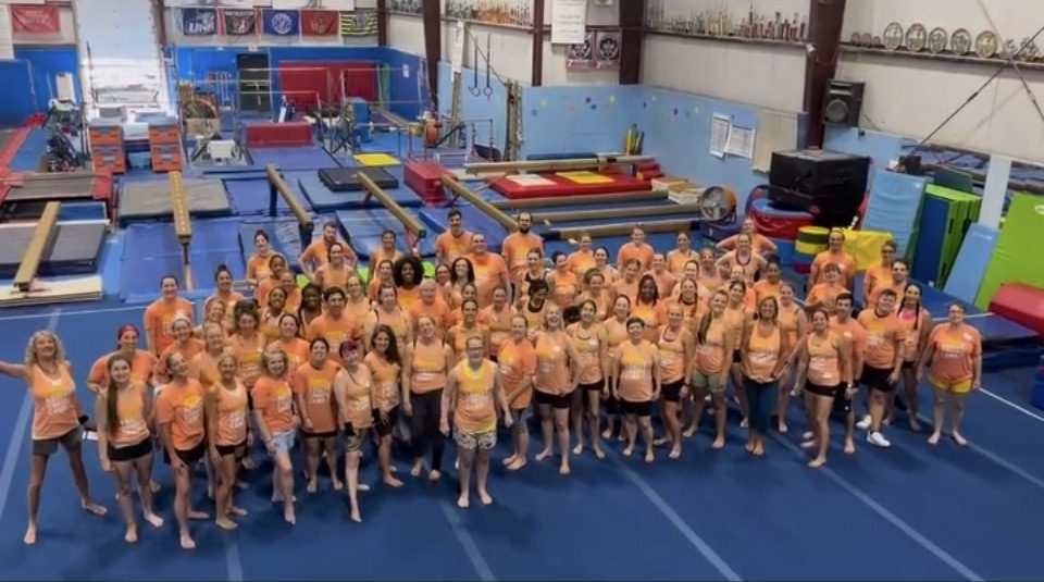 A large group of adult gymnastics camp attendees gathered on the gym floor